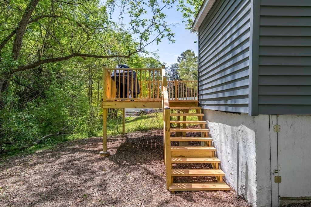 A wooden deck with a staircase leading to a loft area, surrounded by trees and a house.