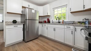 Une cuisine moderne avec des meubles blancs, un réfrigérateur en acier inoxydable, un plan de travail en granit et une fenêtre donnant sur un paysage verdoyant.