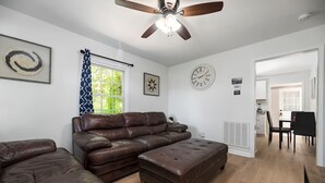 Un salon avec un canapé d’angle en cuir marron, un pouf assorti, un ventilateur de plafond et un coin repas comprenant une table et des chaises.