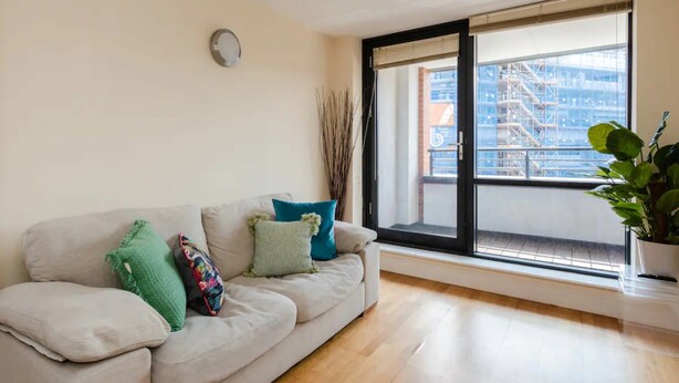 A living room with a sofa, a potted plant, and a large window with a view of a building. - Vivid Apartment in Central London (London)