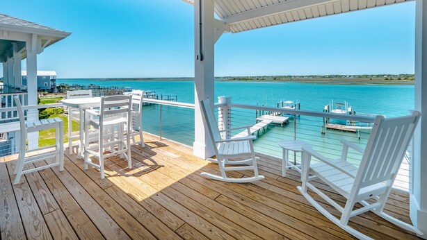 Eine Holzterrasse mit weißen Schaukelstühlen und einem Tisch mit Blick auf eine Marina und Boote.