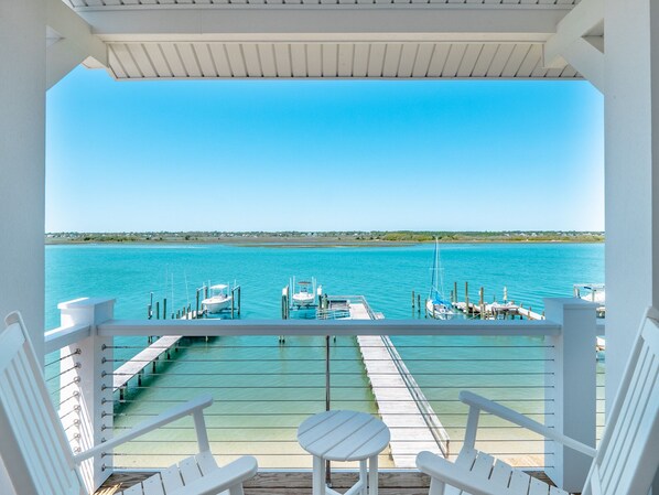 A balcony with white chairs and a small table, overlooking a marina with boats and a clear blue sky.