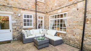 A stone-walled courtyard with a white sofa and wicker ottoman, two windows, and a door.