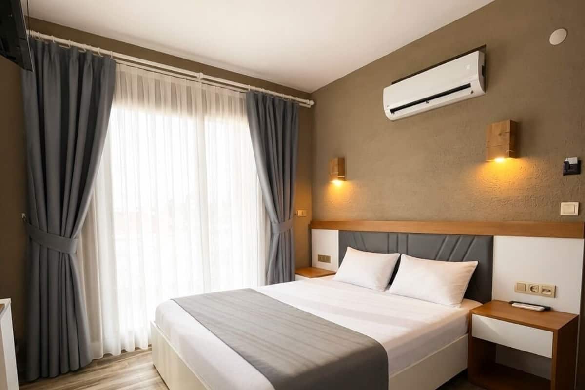 A hotel room with a bed, bedside tables, an air conditioning unit, a window with curtains, and a wooden floor.
