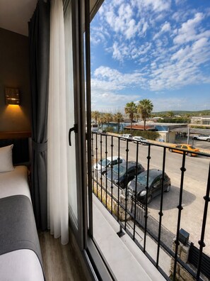 A hotel room with a bed, a window with a view of parked cars and buildings, and a balcony railing. - AURORA SIĞACIK HOTEL (Seferihisar)