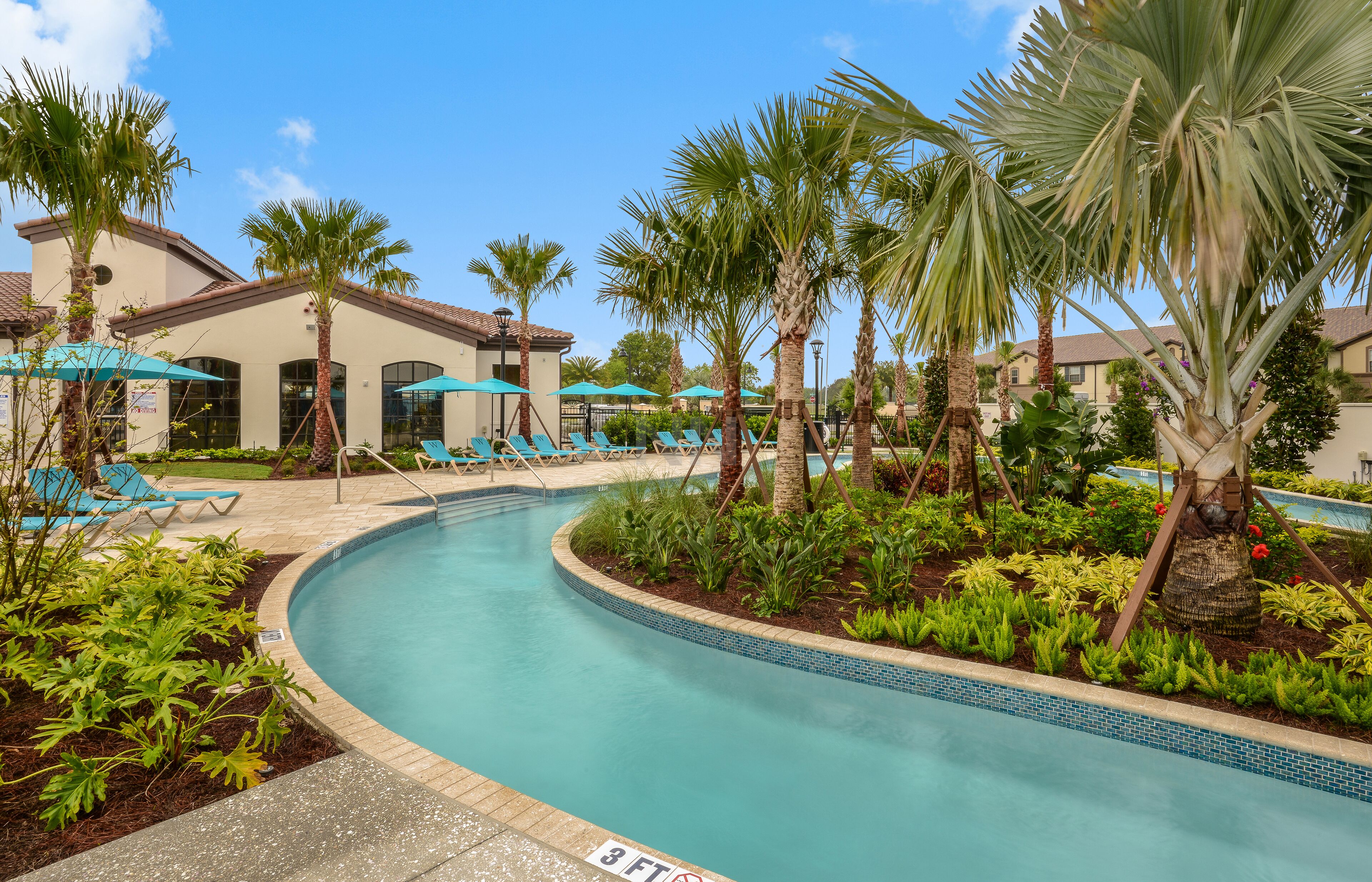 A pool area with lounge chairs and umbrellas, surrounded by palm trees and a building with a tiled roof.
