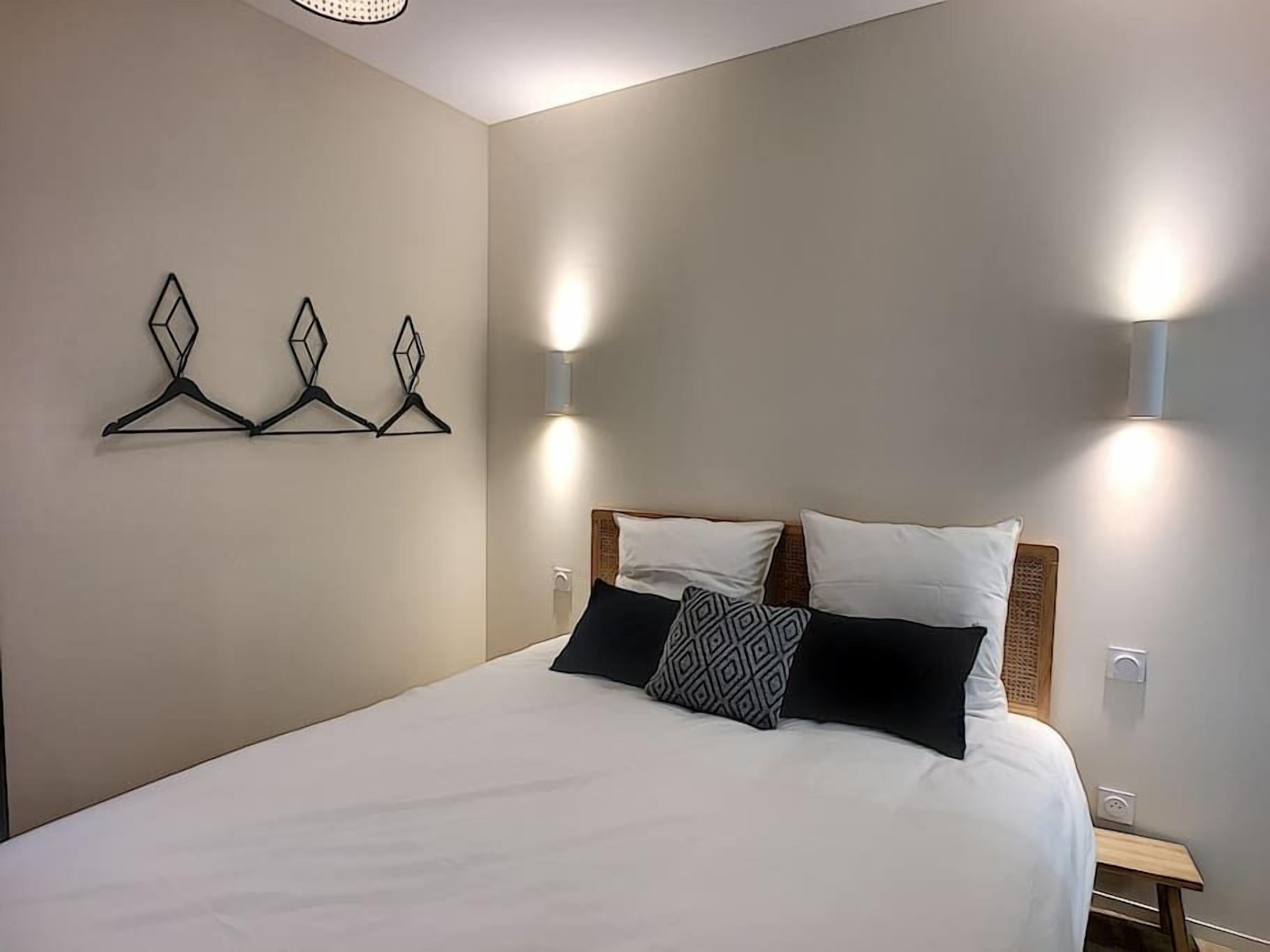 A neatly made bed with a white comforter and two pillows, a wall-mounted light fixture, and a simple bedside table.