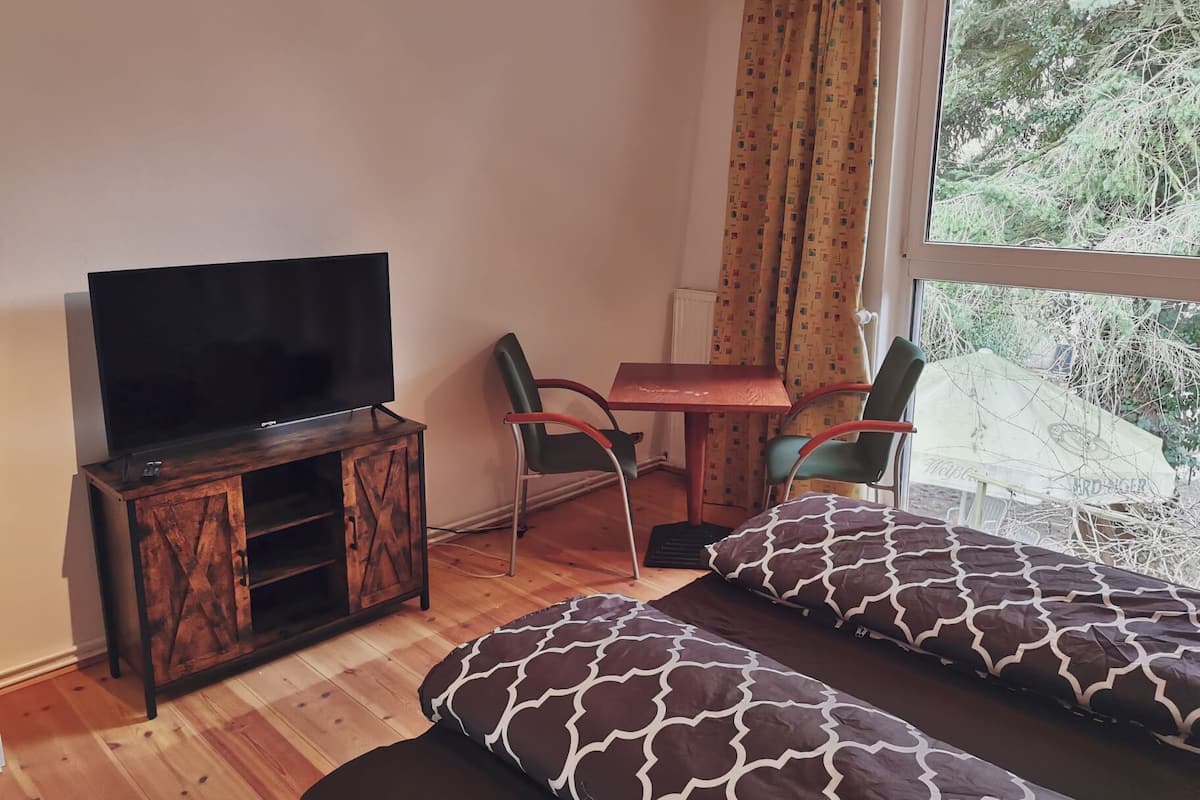 Ein Zimmer mit Fernseher auf einer Holzkommode, einem kleinen Tisch, zwei Stühlen und einem Fenster mit Blick auf Bäume.