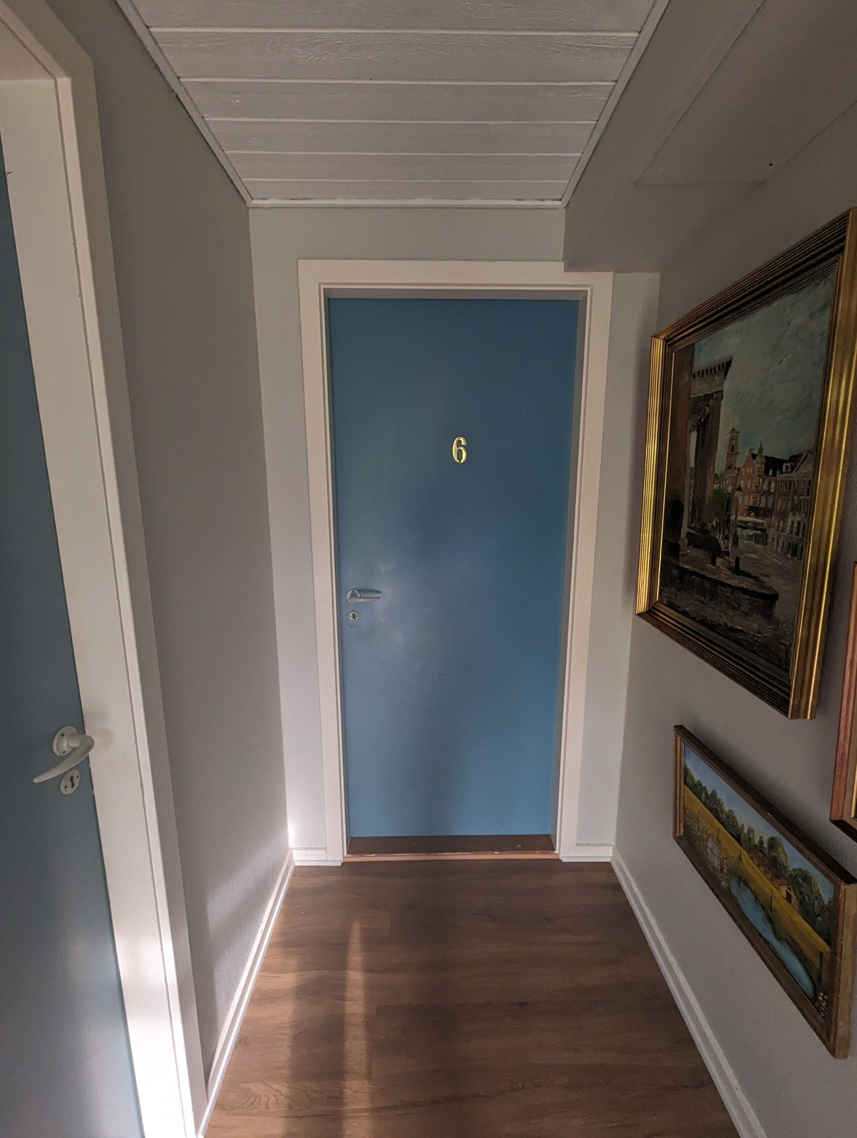 Un passadís amb una porta blava numerada amb el número 6, terres de fusta i obra emmarcada a les parets.