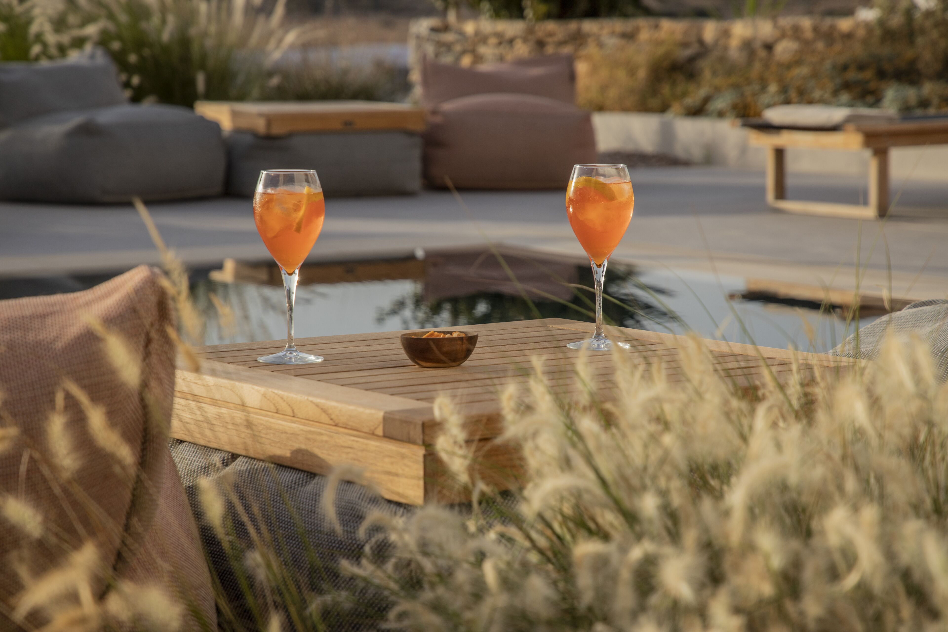Two glasses of drink on a wooden table with a bowl in the center, surrounded by outdoor seating and a pool.