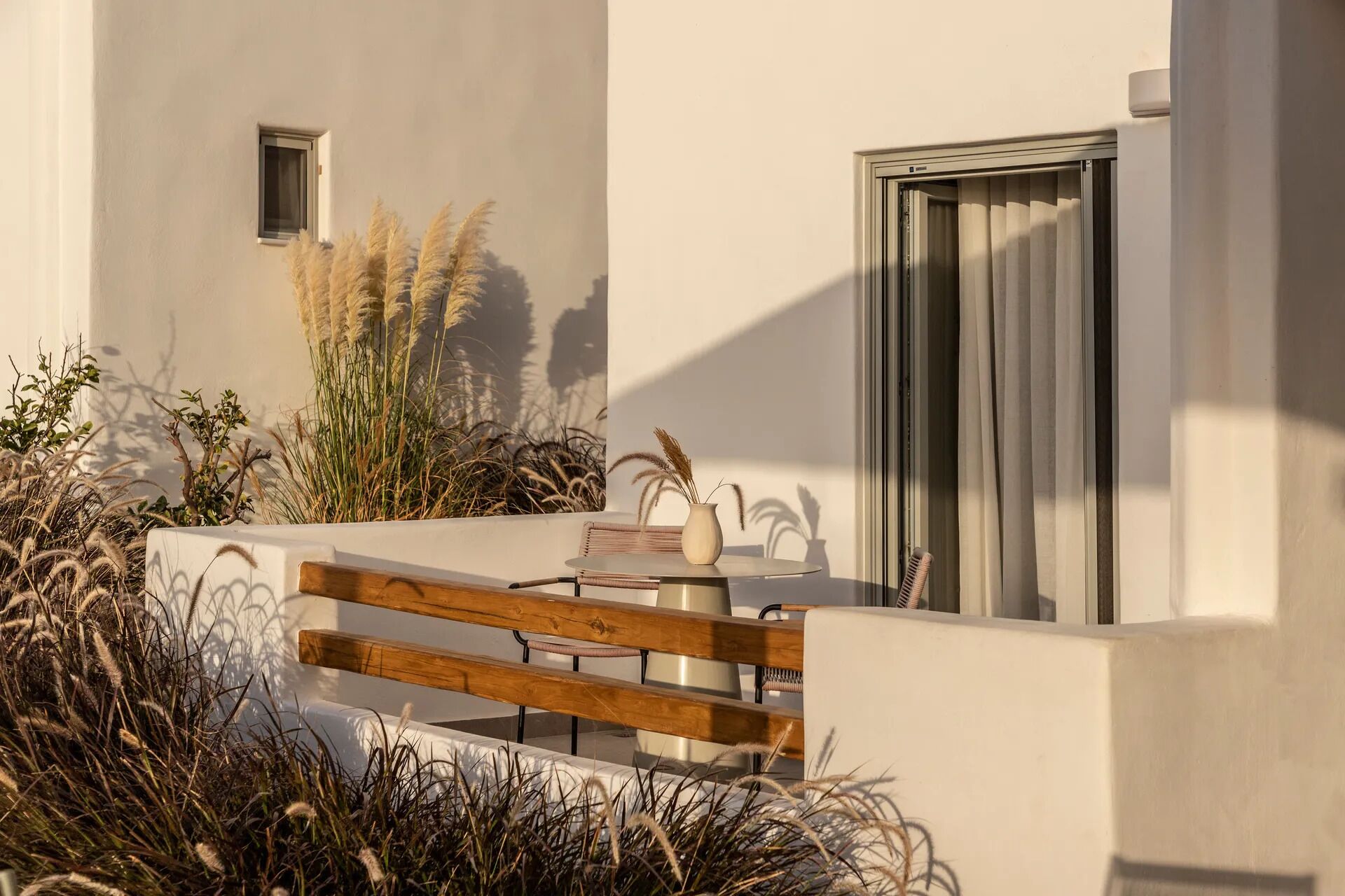A modern outdoor seating area with a wooden bench, a small table, and a vase of pampas grass.