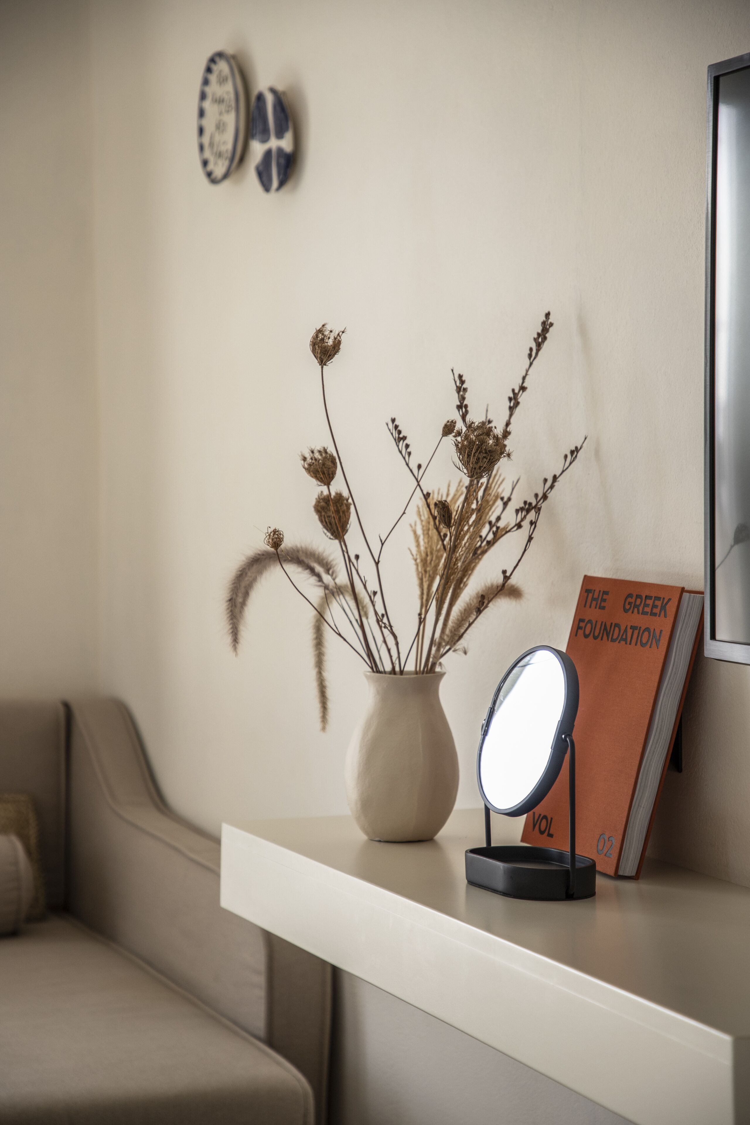 A modern interior with a white shelf holding a vase with dried plants, a book titled "The Greek Foundation," and a round mirror.