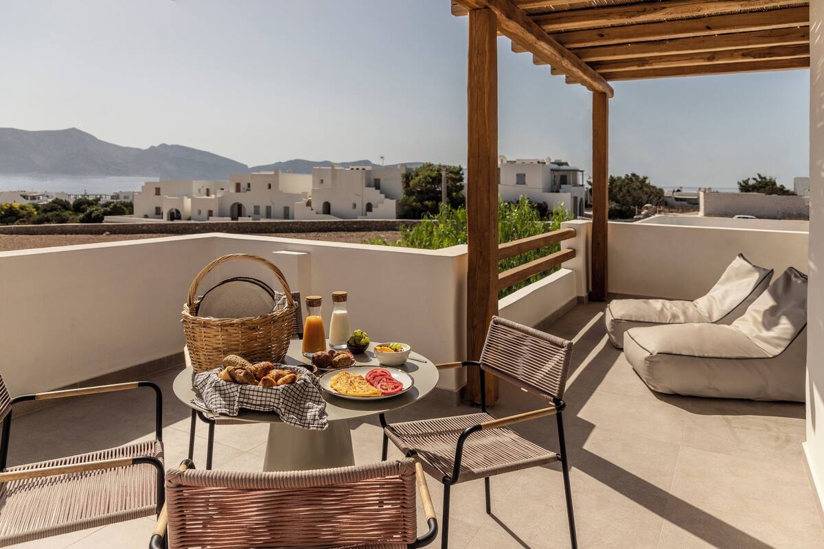 A terrace with wicker chairs, a table with breakfast, and a view of buildings and mountains.