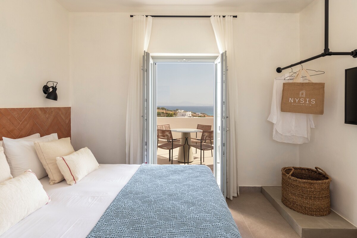A hotel room with a bed, a view of the sea, a table and chairs outside, and a hanging bag with the hotel's name.
