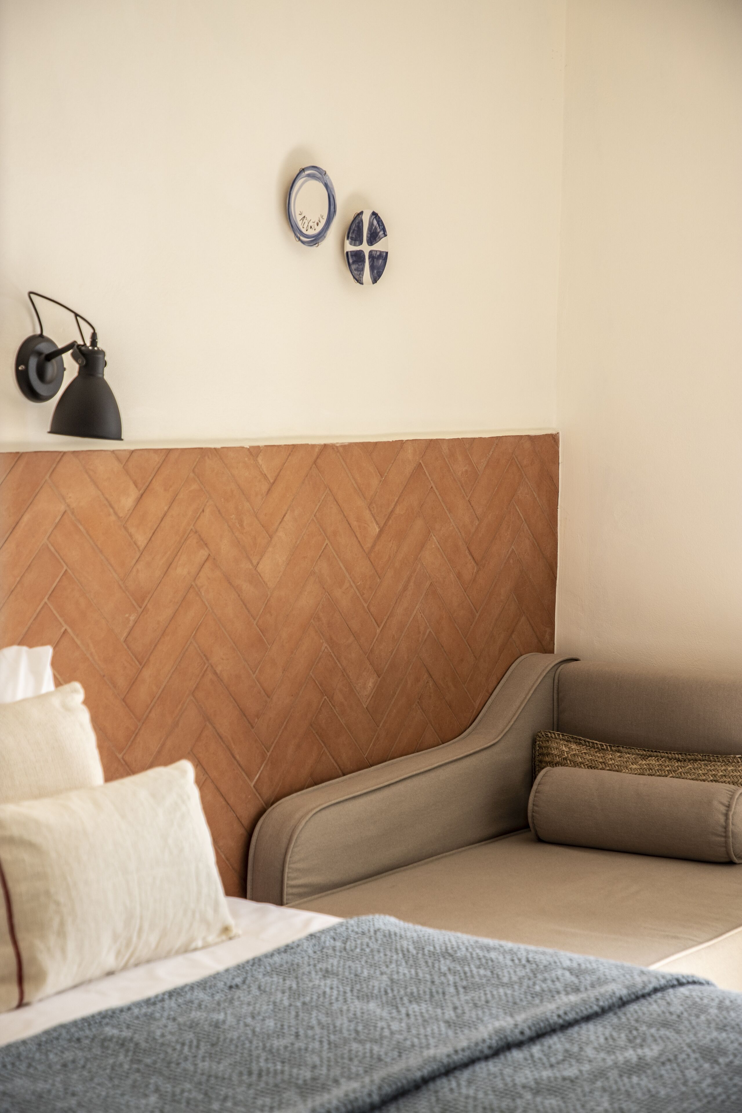 A bedroom with a herringbone-patterned headboard, a bed with white and gray bedding, and a small bedside table with a lamp.