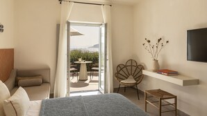 A bedroom with a bed, a chair, a small table, a TV, and a view of the outdoors. - Nysis Koufonisia (Naxos)