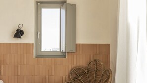 A bathroom with a wicker chair, a small table with a pitcher and glass, and a window with a towel hanging on the wall. - Nysis Koufonisia (Naxos)