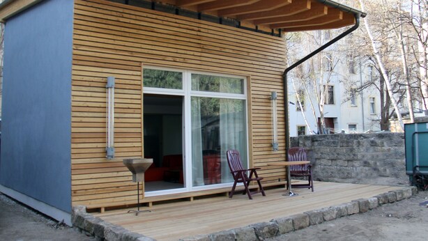 Una cabaña moderna de madera con un amplio ventanal corredera, una pequeña terraza con dos sillas y una pared de piedra al fondo.