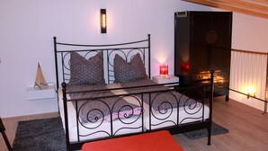 Una cama con estructura metálica, dos almohadas, una mesita de noche, un cojín rojo, una lámpara de pared y un calefactor.