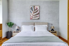 A neatly made bed with white linens, two bedside tables with lamps, a potted plant, and a wall-mounted artwork.
