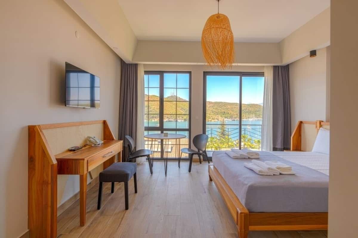 A hotel room with a bed, desk, chairs, a TV, and large windows offering a view of the outdoors.