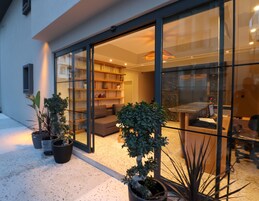 Una casa moderna con un puerta de vidrio que da a una sala de estar con un sofá y una mesa de comedor. Hay plantas en macetas afuera.