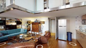 Un espacio habitable amplio con un sofá azul, un sillón de cuero marrón y una mesa de centro de madera. Se ve una cocina con un refrigerador blanco y salpicadero con diseño.