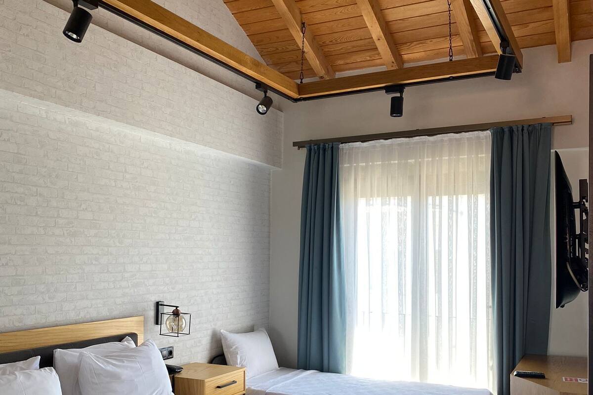 Habitación de hotel con dos camas, vigas de techo de madera, pared de ladrillo y un ventanal con cortinas.