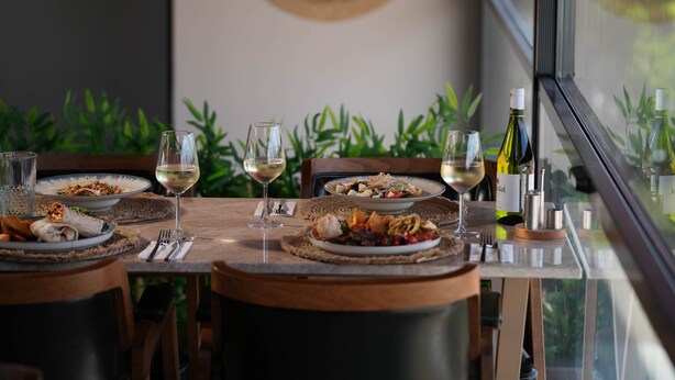Een eettafel gedekt met borden eten, wijnglazen en een fles wijn, met uitzicht op groen door een raam.