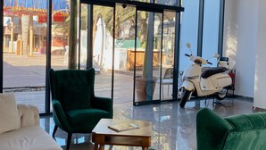 Een moderne kamer met een bank, een groene fauteuil en een kleine tafel. Een witte scooter staat buiten het raam geparkeerd.