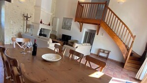 A spacious dining area with a wooden table and chairs, a fireplace, and a staircase leading to an upper level. - Le Genet - Ancienne grange avec piscine privé (Chalais)