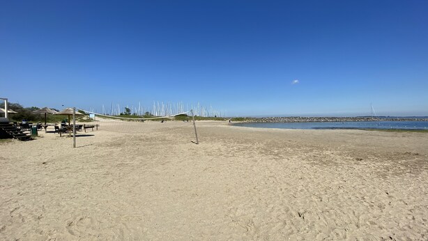 Una playa con una red de voleibol, sombrillas y un muelle al fondo.