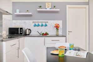 Eine moderne Küche mit Mikrowelle, Wasserkocher und Spüle. Es gibt zwei weiße Schränke, einen weißen Stuhl und einen Tisch mit einem Teller Essen und einem Glas Wasser.