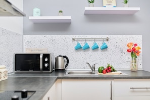A kitchen with a microwave, kettle, and hanging mugs. There are shelves above the counter with plants and a box labeled 'TEA TIME.'