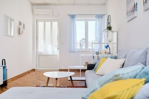 Una sala de estar con un sofá, una mesita redonda, una puerta y una ventana con cortinas.