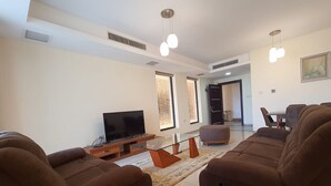 A modern living room with a flat-screen TV, brown sofas, a glass coffee table, and pendant lights. - Abdoun Panorama (Amman)