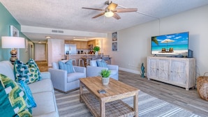 A living room with a sofa, coffee table, and TV, a kitchen in the background, and a ceiling fan. - DIRECT BEACHFRONT! SWIMMING POOL! (Indian Shores)
