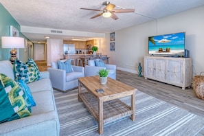 A living room with a sofa, coffee table, and TV, a kitchen in the background, and a ceiling fan.