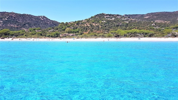 Una playa costera con aguas turquesas cristalinas, una orilla arenosa y edificios en una colina al fondo.