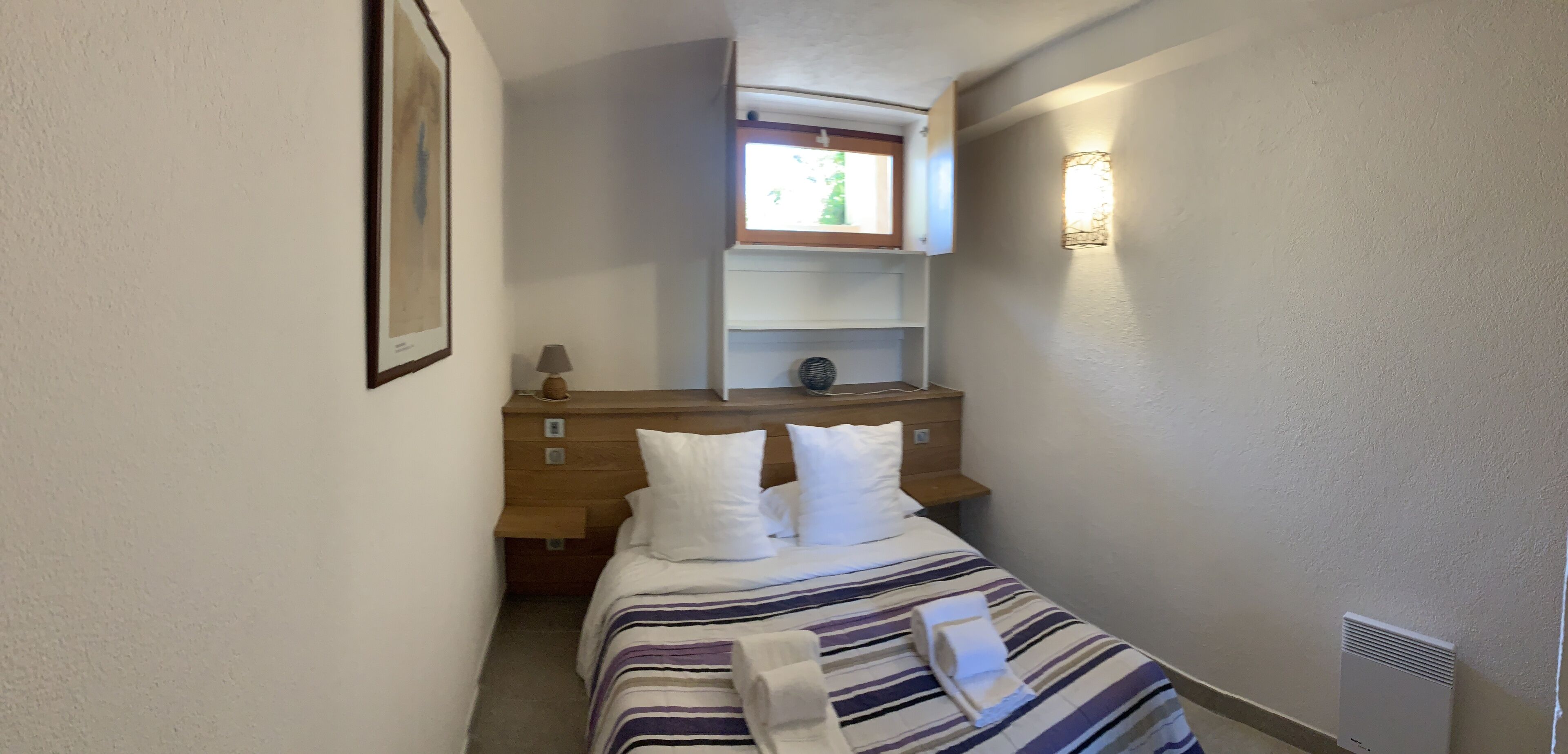 A narrow hotel room with a bed, a nightstand, a lamp, a window, and a wall-mounted shelf.