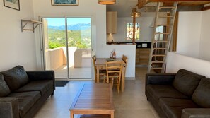 Una sala de estar con dos sofás, una mesa de centro de madera y una mesa de comedor con sillas. Hay un ventanal corredizo que da a un balcón con vistas a las montañas. Se ve una cocina al fondo.