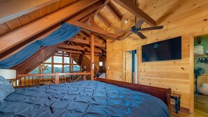Une chambre avec un grand lit, des poutres apparentes en bois au plafond, un ventilateur de plafond et un téléviseur fixé au mur.