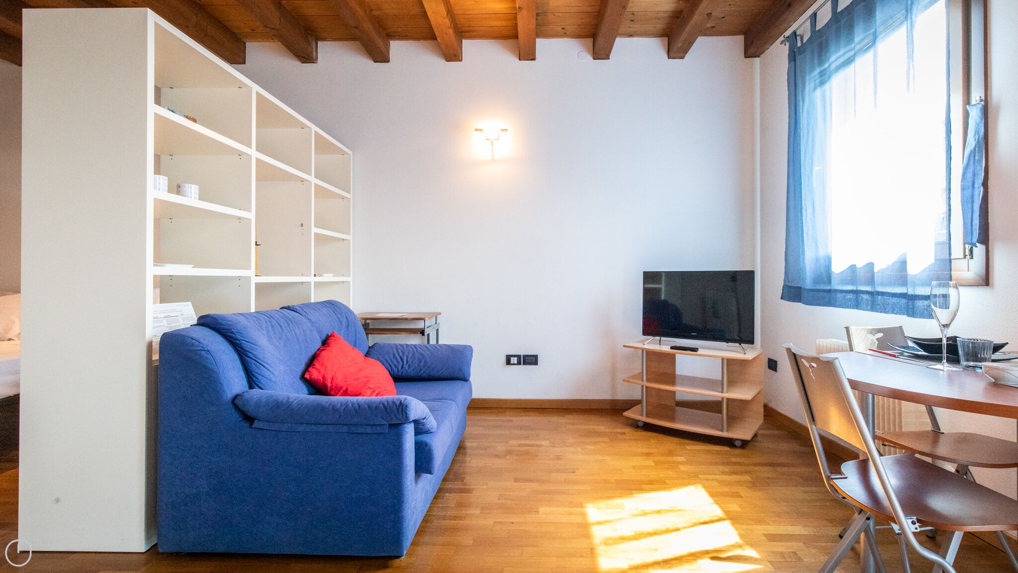 Ein kompakter Wohnraum mit einem blauen Sofa, einem Fernseher auf einem Schrank, einem Esstisch und einem Fenster mit Vorhängen.