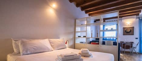 A bedroom with a bed, a bedside table, a shelf unit, and a desk with a chair.