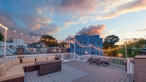 Una terraza en la azotea con mobiliario exterior, luces de hilo y vistas de edificios circundantes.