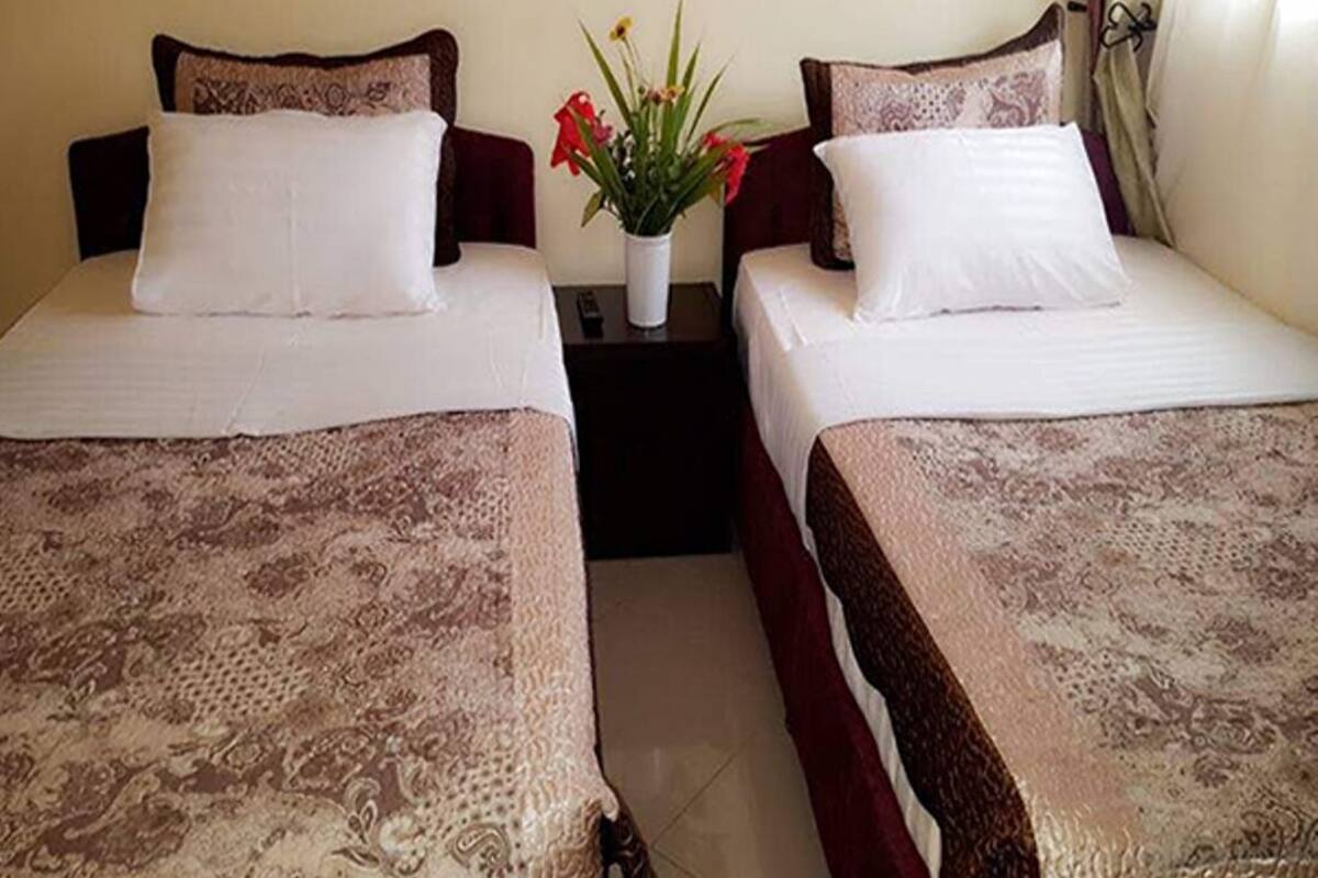 Habitación de hotel con dos camas, cada una con una colcha estampada y almohadas a juego. Entre las camas, hay una mesita de noche con un jarrón de flores.