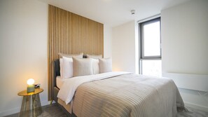 A modern bedroom with a bed, a bedside lamp, a window, and a wooden panel wall.