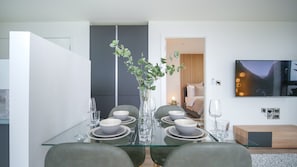 A modern dining area with a glass table, grey chairs, and a vase with green foliage.