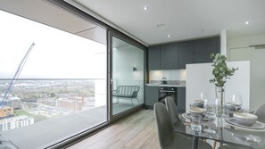 A modern kitchen with a glass door leading to a balcony, a dining area with a table set for two, and a view of a construction site.