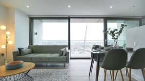 A modern living room with a sofa, dining table, and chairs, a large window with a view of the city, and a balcony.
